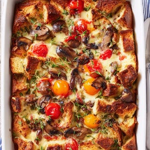 Può includere: Una casseruola al forno in una pirofila rettangolare bianca. Il piatto contiene pane a cubetti, funghi, pomodorini rossi e gialli ed erbe aromatiche. Un cucchiaio poggia su un panno a righe blu e bianche.