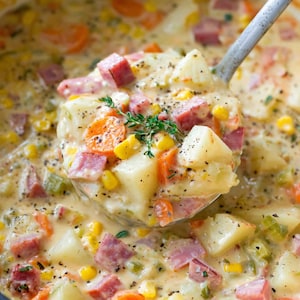 May include: A creamy soup with visible chunks of potatoes, carrots, corn, and ham. A ladle scoops up a portion of the soup, showcasing the ingredients. The soup is in a dark pot, and the background is slightly blurred.