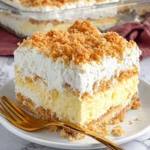 May include: A slice of layered dessert on a white plate with a gold fork. The dessert features a crumbly base, a creamy yellow layer, a white whipped topping, and a crunchy, golden topping. A larger dish of the same dessert is in the background.