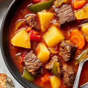以下が含まれることがあります： 黒いボウルに入った、牛肉、ジャガイモ、ニンジン、ピーマン、トマトを濃厚な赤いスープで煮込んだ、ボリュームたっぷりのビーフシチューのクローズアップ。銀のスプーンがシチューの中にあり、カリカリのパンのスライスが横にあります。