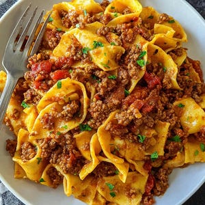 May include: A plate of pasta with a meat sauce. The pasta is a wide, flat shape, and the sauce is a rich brown color with visible pieces of tomato and herbs. A silver fork rests on the side of the plate. The dish is on a white plate.