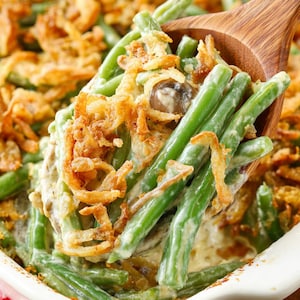 May include: A close-up of a casserole dish filled with green beans, a creamy sauce, and crispy fried onions. A wooden spoon lifts a portion of the dish, highlighting the textures and colours of the food.