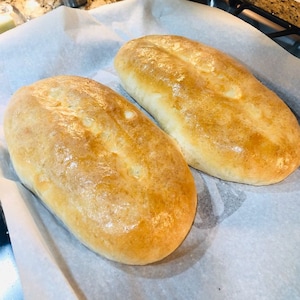 May include: Two golden-brown, freshly baked loaves of bread rest on parchment paper. The bread has a glossy sheen, indicating it's just out of the oven. The loaves are oblong, with slight indentations on top, suggesting a soft interior. A culinary image.