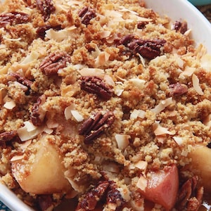 Puede incluir: Crocante de manzana horneado en una fuente ovalada blanca. El postre presenta una cobertura dorada y desmenuzable con nueces pecanas y copos de coco. Las manzanas son visibles debajo de la cobertura.