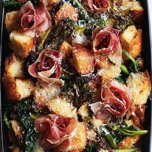 May include: A baked dish featuring golden-brown bread cubes, dark green leafy vegetables, and delicate prosciutto roses. The dish is sprinkled with grated cheese and presented in a black baking pan, showcasing a rustic and appetizing meal.