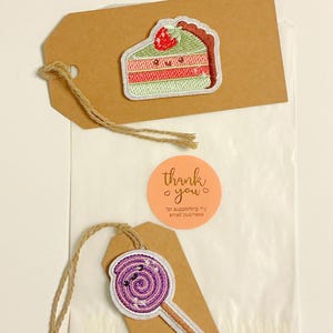 May include: A brown tag with a slice of cake patch and a purple lollipop patch. The cake patch has green, pink, and red colors. The lollipop patch is purple with white accents. A round sticker with the words "thank you" is also visible.
