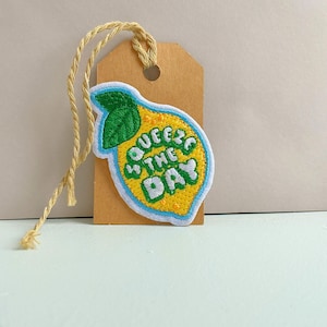May include: A yellow lemon-shaped patch with the text "Squeeze the Day" in white and green, outlined in blue. The patch is attached to a brown tag with a natural twine string.