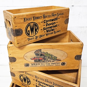 May include: Stack of three wooden crates with a vintage Great Western Railway (GWR) logo and text. The top crate has the GWR logo and text "DAILY DIRECT ROUTES FROM LONDON - Cornwall - Devon - Somerset - Wales - TRAVEL BY GREAT WESTERN RAILWAYS". The middle crate has a vintage illustration of a train with the text "SPEED TO THE WEST GWR CORNWALL DEVON SOMERSET WALES". The bottom crate has the GWR logo and text "DAILY DIRECT ROUTES FROM LONDON - Cornwall - Devon - Somerset - TRAVEL BY GREAT WESTERN RAILWAYS".
