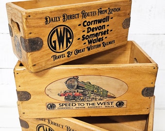 May include: Stack of three wooden crates with a vintage Great Western Railway (GWR) logo and text. The top crate has the GWR logo and text "DAILY DIRECT ROUTES FROM LONDON - Cornwall - Devon - Somerset - Wales - TRAVEL BY GREAT WESTERN RAILWAYS". The middle crate has a vintage illustration of a train with the text "SPEED TO THE WEST GWR CORNWALL DEVON SOMERSET WALES". The bottom crate has the GWR logo and text "DAILY DIRECT ROUTES FROM LONDON - Cornwall - Devon - Somerset - TRAVEL BY GREAT WESTERN RAILWAYS".