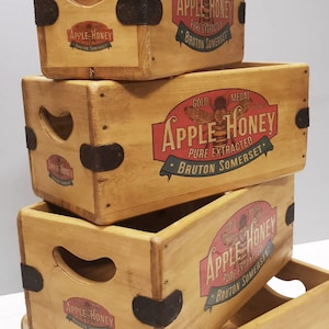 May include: Four stacked wooden crates, each bearing the words "Apple Honey" in red and blue. The crates are light brown, with dark metal corner protectors. The text also includes "Bruton Somerset".