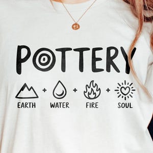 May include: Cream-colored t-shirt with the word "POTTERY" in bold black letters. Below the word are four symbols representing earth, water, fire, and soul. The shirt is a casual, oversized fit.