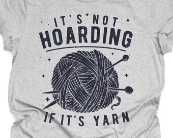 Op de afbeelding: Grijs T-shirt met een blauwe afbeelding van een bol garen en breinaalden. De tekst op het shirt luidt "It's not hoarding if it's yarn."