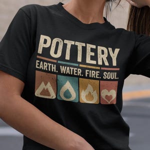 May include: Black t-shirt with the word "POTTERY" in a vintage font, with the words "EARTH. WATER. FIRE. SOUL." below. The shirt features four small, colourful graphic icons. The model is wearing blue jeans.