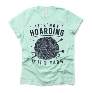 May include: A light green t-shirt with a black and white graphic of a ball of yarn and knitting needles. The text on the shirt reads "It's not hoarding if it's yarn."