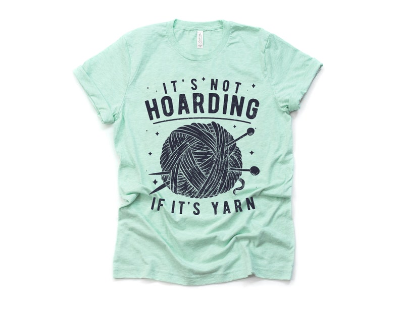 May include: A light green t-shirt with a black and white graphic of a ball of yarn and knitting needles. The text on the shirt reads "It's not hoarding if it's yarn."