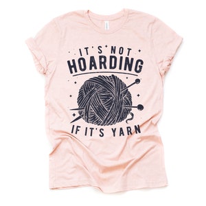 May include: A pink t-shirt with a black and white graphic of a ball of yarn and knitting needles. The text on the shirt reads "It's not hoarding if it's yarn."