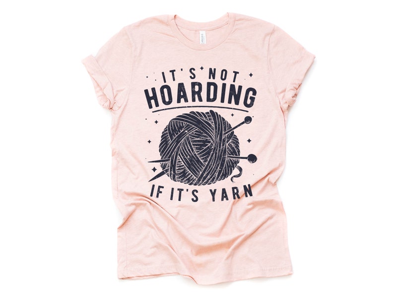 May include: A pink t-shirt with a black and white graphic of a ball of yarn and knitting needles. The text on the shirt reads "It's not hoarding if it's yarn."