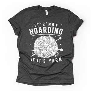 May include: A dark gray t-shirt with a white graphic of a ball of yarn and knitting needles. The text "It's not hoarding if it's yarn" is printed on the shirt.