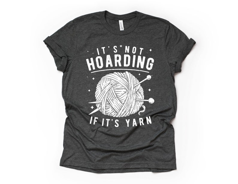 May include: A dark gray t-shirt with a white graphic of a ball of yarn and knitting needles. The text "It's not hoarding if it's yarn" is printed on the shirt.