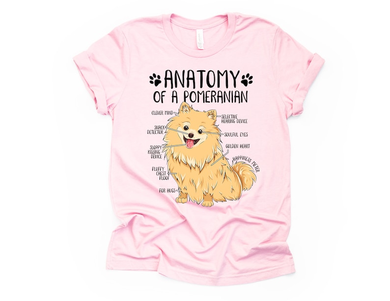Op de afbeelding: Roze T-shirt met een cartoon Pomeranian hond en de tekst "Anatomie van een Pomeranian" met humoristische beschrijvingen van de kenmerken van de hond.