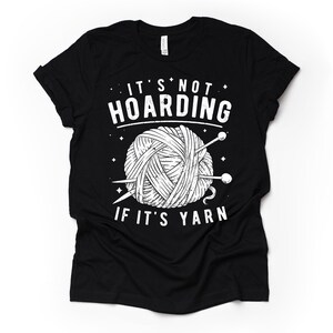 May include: Black t-shirt with a white graphic of a ball of yarn and knitting needles. The text "It's not hoarding if it's yarn" is printed on the shirt.