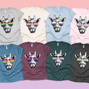 Pode incluir: Uma coleção de camisetas em várias cores, cada uma com um gráfico de uma girafa usando óculos de sol com diferentes designs de bandeiras do orgulho. As camisas são de manga curta e feitas de um material macio.