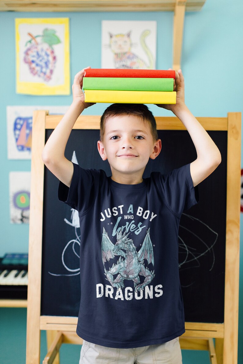 May include: A young boy wearing a navy blue t-shirt with a graphic of a dragon and the text "Just a boy who loves dragons". He is holding a stack of three colorful books above his head.