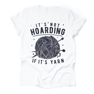 May include: White t-shirt with a black and white graphic of a ball of yarn and knitting needles. The text "It's not hoarding if it's yarn" is printed on the shirt.