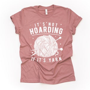 May include: A mauve t-shirt with a white graphic of a ball of yarn and knitting needles. The text on the shirt reads "It's not hoarding if it's yarn."
