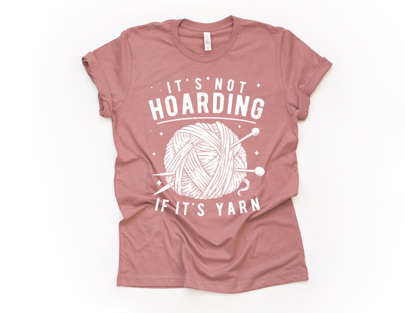 May include: A mauve t-shirt with a white graphic of a ball of yarn and knitting needles. The text on the shirt reads "It's not hoarding if it's yarn."