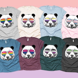 Puede incluir: Una colección de camisetas con un panda que lleva gafas de sol con varios diseños de banderas del orgullo LGBTQ+. Las camisetas están disponibles en una variedad de colores, incluyendo gris, crema, azul y rosa. El gráfico del panda es en blanco y negro.