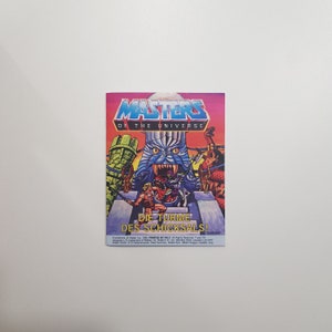 Puede incluir: Una carta de colección de Masters of the Universe con He-Man y Skeletor. La carta tiene una ilustración colorida de los dos personajes luchando frente a un castillo. El texto en la carta dice "Die Turme Des Schicksals!"