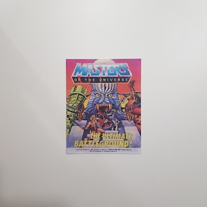 Puede incluir: Una tarjeta de intercambio vintage de Masters of the Universe con He-Man y Skeletor luchando en un fondo colorido y de fantasía. La tarjeta dice "The Ultimate Battleground!"