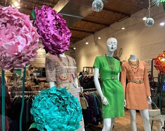 Könnte beinhalten: Drei Schaufensterpuppen in einem Schaufenster, die Vintage-Kleider tragen. Die Schaufensterpuppen sind mit großen Papierblumen in Pink, Türkis und Weiß verziert. Das Schaufenster ist mit Discokugeln verziert, die von der Decke hängen.