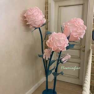 May include: A large, decorative floral arrangement featuring oversized pink paper roses. The roses are mounted on teal metal stems with green leaves. The arrangement is placed indoors near a door.