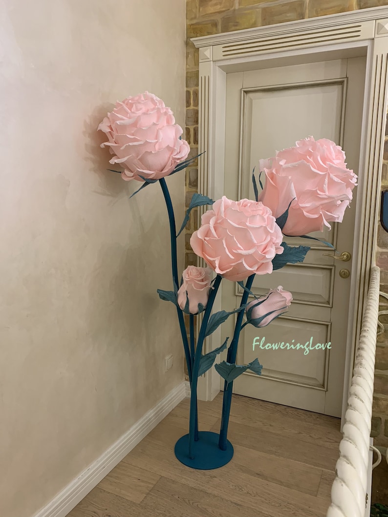 May include: A large, decorative floral arrangement featuring oversized pink paper roses. The roses are mounted on teal metal stems with green leaves. The arrangement is placed indoors near a door.