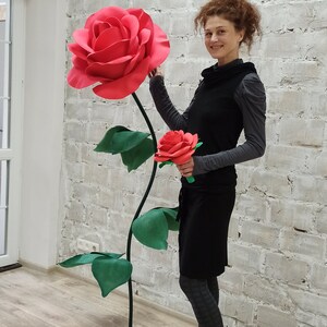 May include: A large, red paper rose with green leaves and a long stem stands next to a smaller red rose bouquet. The large rose is approximately 5 feet tall. The background is a white brick wall.