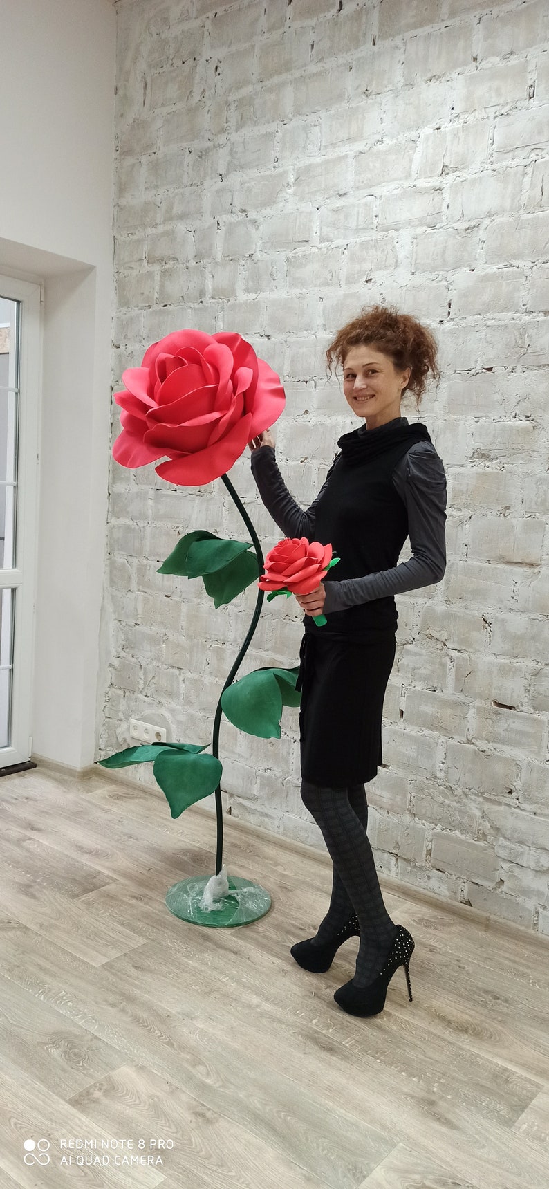 May include: A large, red paper rose with green leaves and a long stem stands next to a smaller red rose bouquet. The large rose is approximately 5 feet tall. The background is a white brick wall.