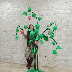 May include: A whimsical, handcrafted apple tree sculpture. The tree features a green base and stems, with large green leaves and pink apples. The tree is made of felt and wire, and stands approximately 6 feet tall.