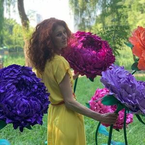 K&ouml;nnte beinhalten: Eine Frau in einem gelben Kleid steht auf einem Feld mit riesigen Papierblumen. Die Blumen sind lila, rosa und blau. Die Frau l&auml;chelt und schaut in die Kamera.