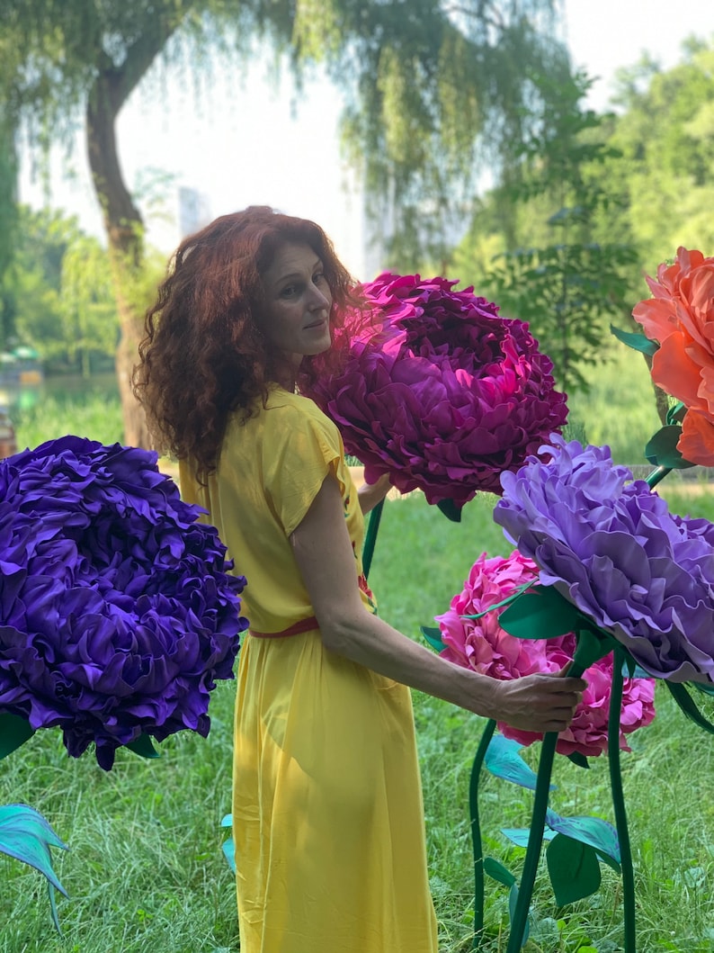 K&ouml;nnte beinhalten: Eine Frau in einem gelben Kleid steht auf einem Feld mit riesigen Papierblumen. Die Blumen sind lila, rosa und blau. Die Frau l&auml;chelt und schaut in die Kamera.