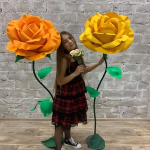 May include: A young person stands with two large, artificial roses. One rose is orange, the other is yellow. The roses have green stems and leaves. The person is wearing a red plaid dress and black sneakers.