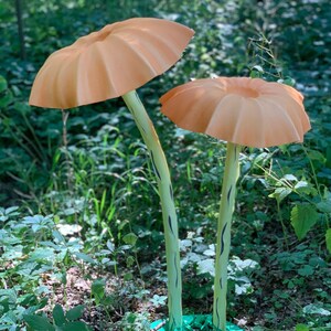 May include: Two large, orange mushroom sculptures with ribbed caps and light green stems with black markings. The mushrooms stand in a lush green forest setting, with a green base at the bottom of the stems.