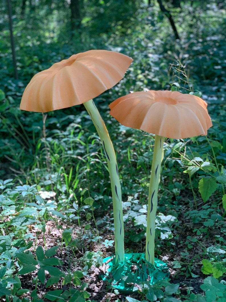 May include: Two large, orange mushroom sculptures with ribbed caps and light green stems with black markings. The mushrooms stand in a lush green forest setting, with a green base at the bottom of the stems.