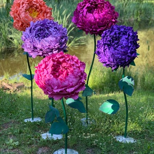 K&ouml;nnte beinhalten: F&uuml;nf gro&szlig;e Papierblumen in verschiedenen Farben sind auf gr&uuml;nem Gras angeordnet. Die Blumen sind rosa, lila und orange. Die Blumen sitzen auf gr&uuml;nen Stielen mit Bl&auml;ttern.