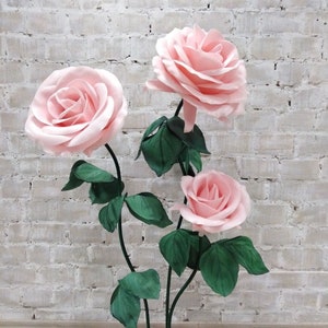 May include: A decorative floral arrangement featuring three large, light pink roses with green leaves and stems. The roses are in full bloom, with detailed petals. The arrangement is set on a green base, creating a striking visual display.