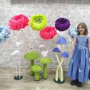 K&ouml;nnte beinhalten: Ein junges M&auml;dchen in einem blauen Kleid steht vor einer Backsteinwand, die mit gro&szlig;en Papierblumen und pilzf&ouml;rmigen Dekorationen geschm&uuml;ckt ist. Die Blumen sind in den Farben Rosa, Blau, Gr&uuml;n und Lila.