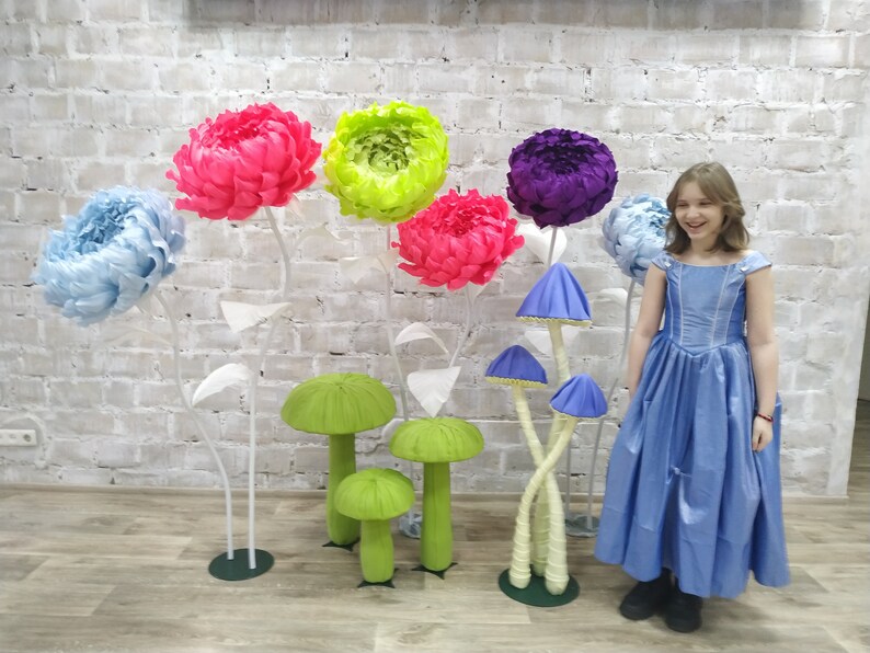 K&ouml;nnte beinhalten: Ein junges M&auml;dchen in einem blauen Kleid steht vor einer Backsteinwand, die mit gro&szlig;en Papierblumen und pilzf&ouml;rmigen Dekorationen geschm&uuml;ckt ist. Die Blumen sind in den Farben Rosa, Blau, Gr&uuml;n und Lila.
