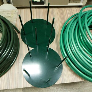 May include: An overhead shot shows a dark green hose and a dark green metal hose holder. The hose is coiled in two separate piles. The hose holder has a circular base with vertical rods. The items are on a light-colored wooden surface.