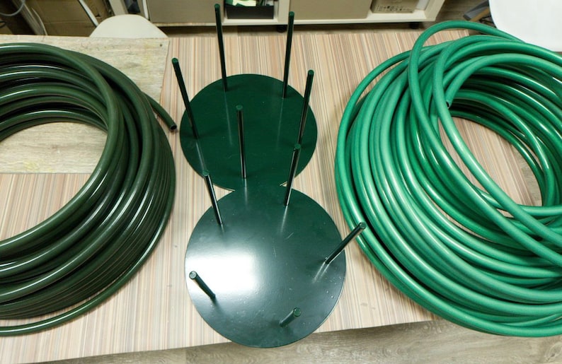 May include: An overhead shot shows a dark green hose and a dark green metal hose holder. The hose is coiled in two separate piles. The hose holder has a circular base with vertical rods. The items are on a light-colored wooden surface.
